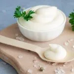 Bowl and spoonful of mayonnaise on a wooden board with fresh parsley