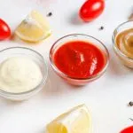 Three glass bowls of white, red and brown dipping sauces with cherry tomatoes, lemon and basil