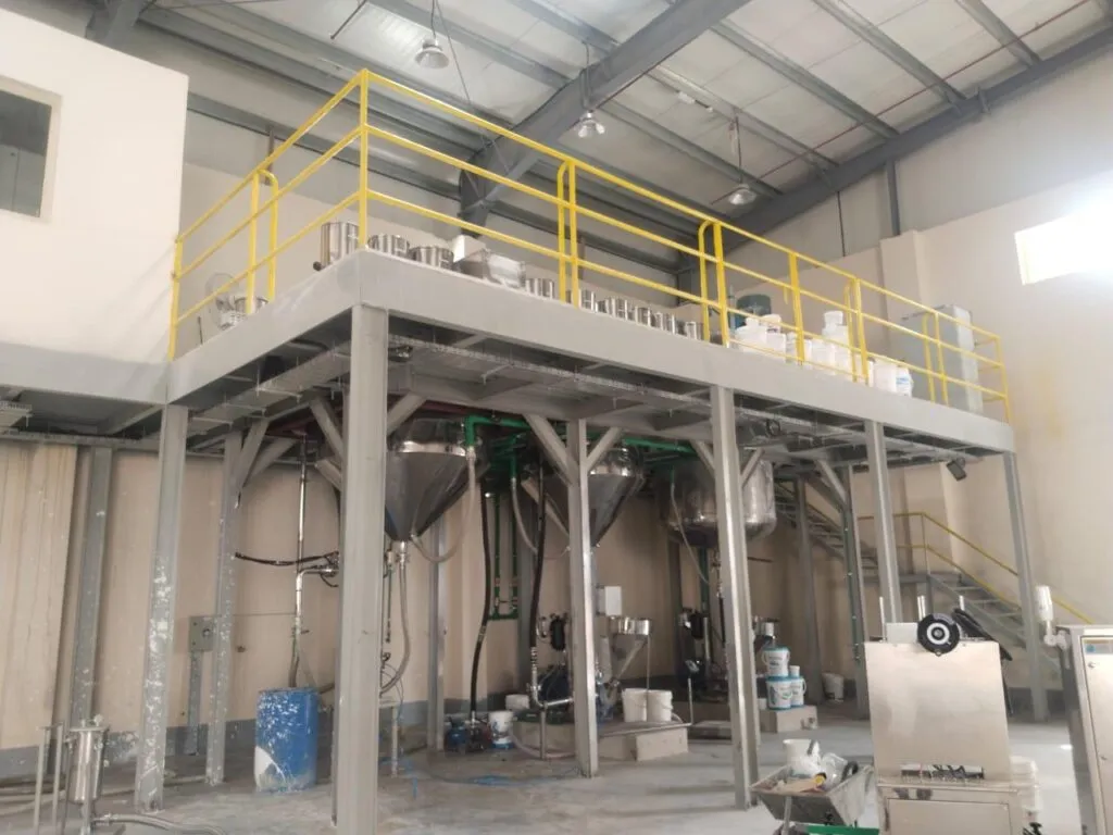 Paint production plant hall with a yellow-railed mezzanine above conical process vessels and paint buckets