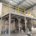 Paint production plant hall with a yellow-railed mezzanine above conical process vessels and paint buckets