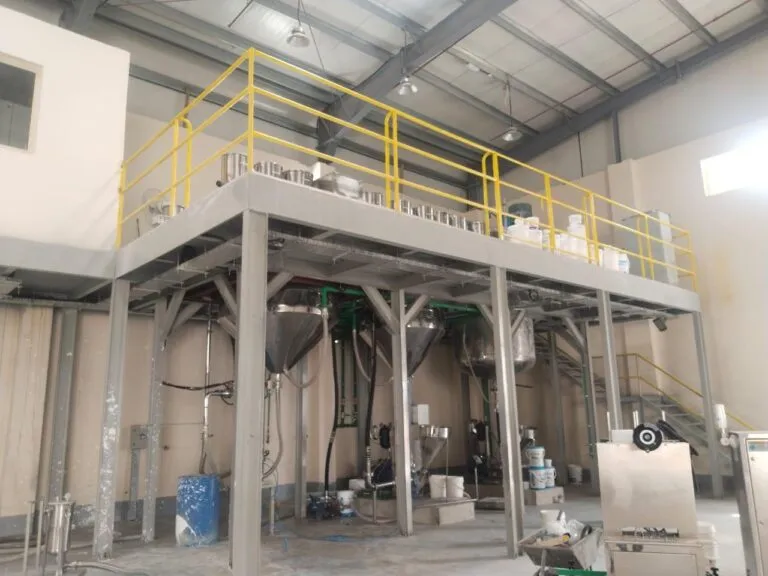 Paint production plant hall with a yellow-railed mezzanine above conical process vessels and paint buckets